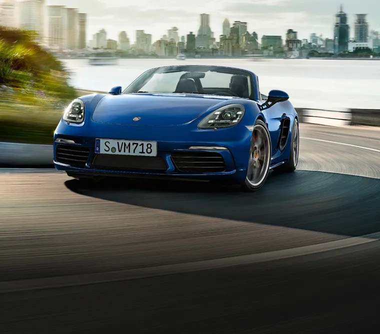 Porsche 718 Boxster S in Gentian Blue Metallic taking a bend on a coastal road. Front and side view.