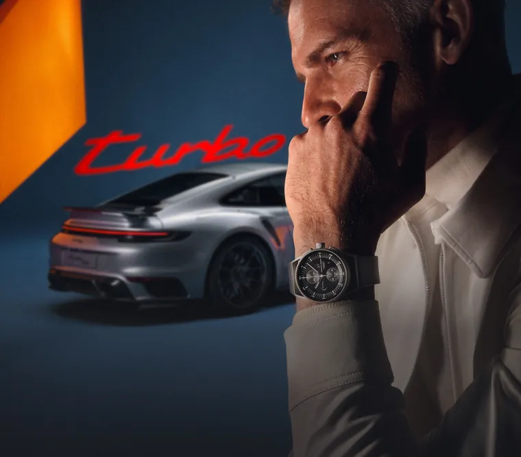 图片展示一位男士腕间佩戴 Porsche Design 911 Turbo 50 周年纪念版腕表站立于一辆 GT 银金属漆保时捷 911 Turbo 50 周年纪念版车型前方 