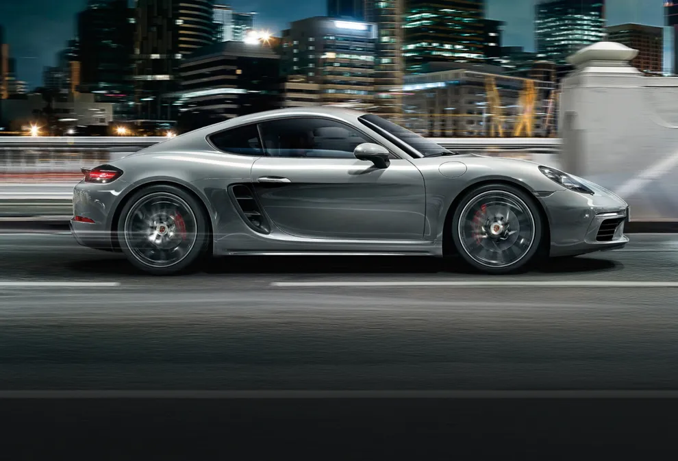 Porsche 718 Cayman S in GT Silver Metallic driving on a road with a city skyline in the background. View of side with flyline.