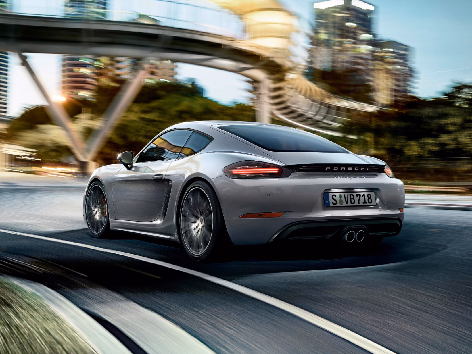 Porsche 718 Cayman S in GT Silver Metallic driving on a road. Rear and side view.