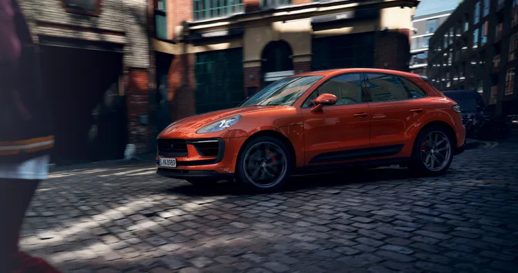 Porsche Macan S in Papaya Metallic driving through the city over cobblestones. Side view.