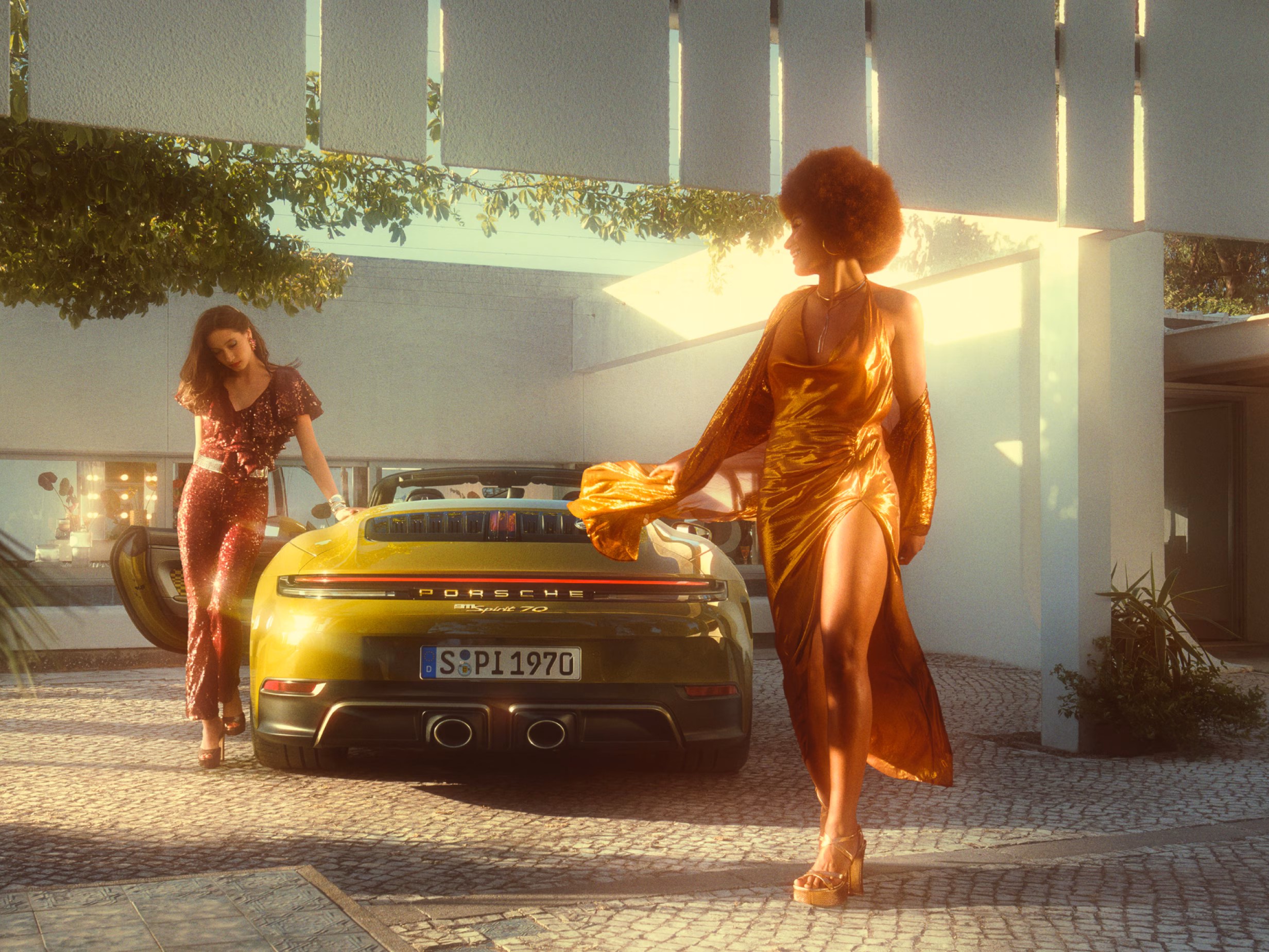 采用橄榄色 Neo 车漆的保时捷 911 Spirit 70 停在铺装广场上。后端后视图两名身着 70 年代服装的女性正从车上下来。