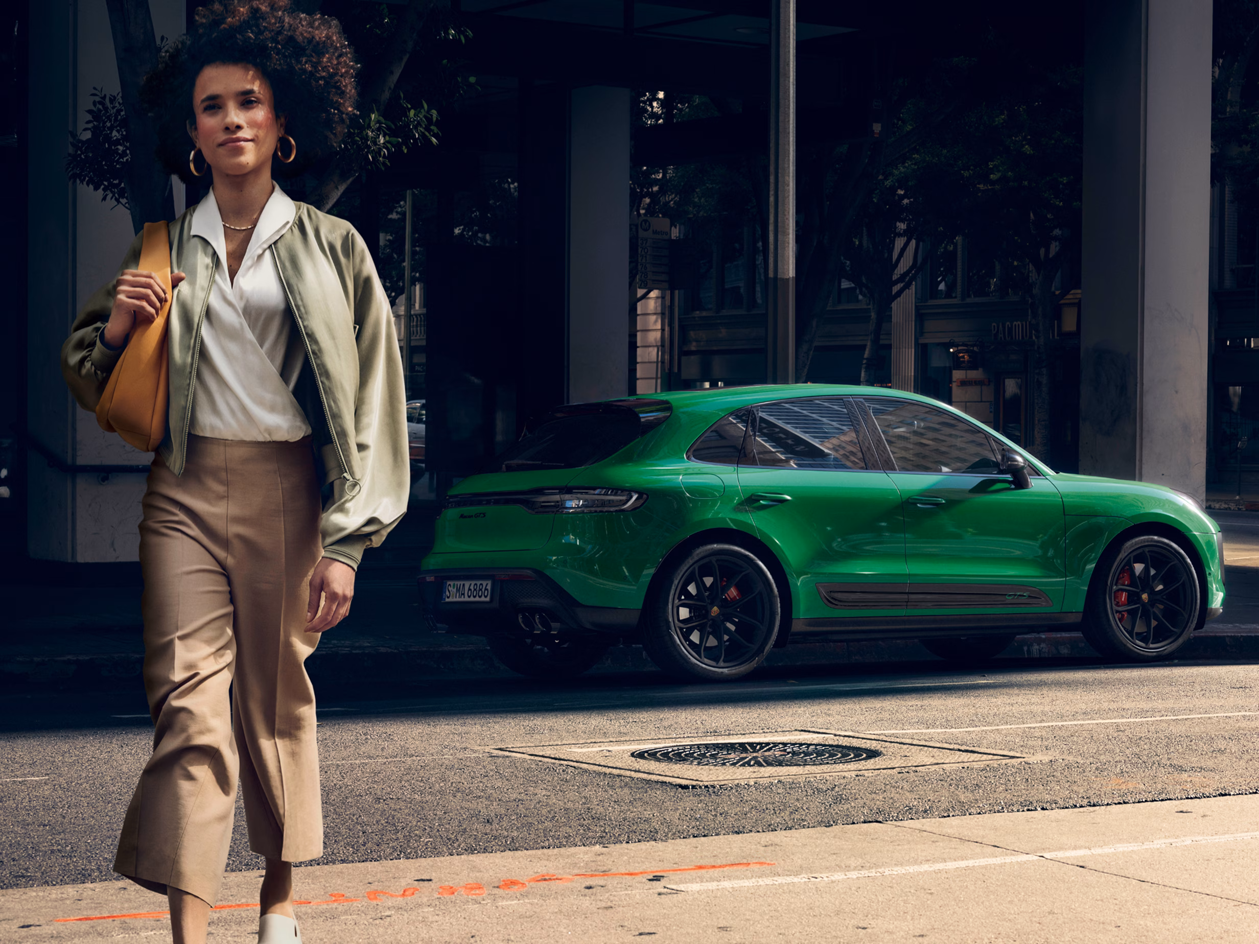 Porsche Macan GTS in Python Green in the city. Rear and side view. A woman is walking past in the foreground.
