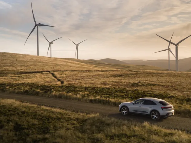 Ein Porsche Macan Turbo in Eisgraumetallic fährt über eine Landstraße durch eine hügelige Graslandschaft. Im Hintergrund stehen Windräder.