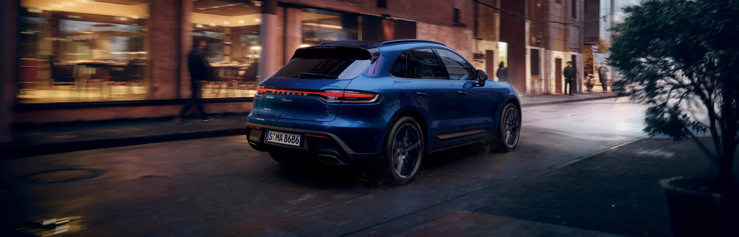 Porsche Macan in Gentian Blue Metallic driving through a city. View of side and rear end.