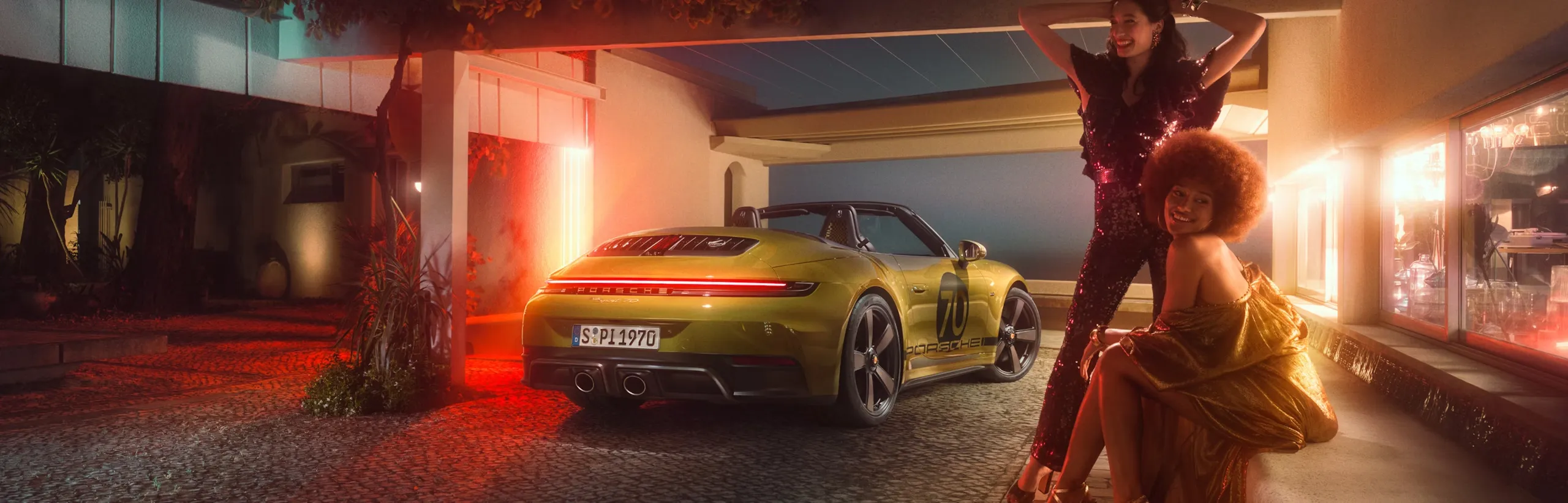 夜间，采用橄榄色 Neo 车漆的保时捷 911 Spirit 70 停在车棚下。从后面看的后侧视图前景中，两名身着 70 年代服装的女性在摆姿势。