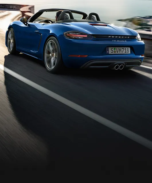 Porsche 718 Boxster S in Gentian Blue Metallic driving along a coastal road. View of the rear and the side.