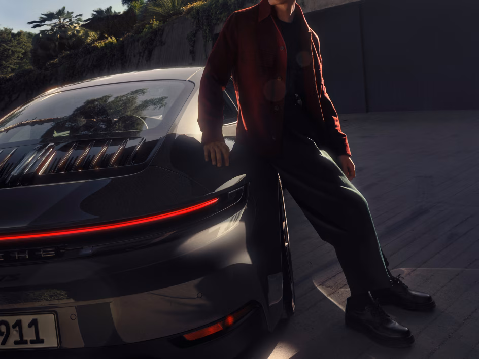 Man leaning against the side of the Porsche 911 Carrera GTS in Slate Grey Neo shading his face from the sun.