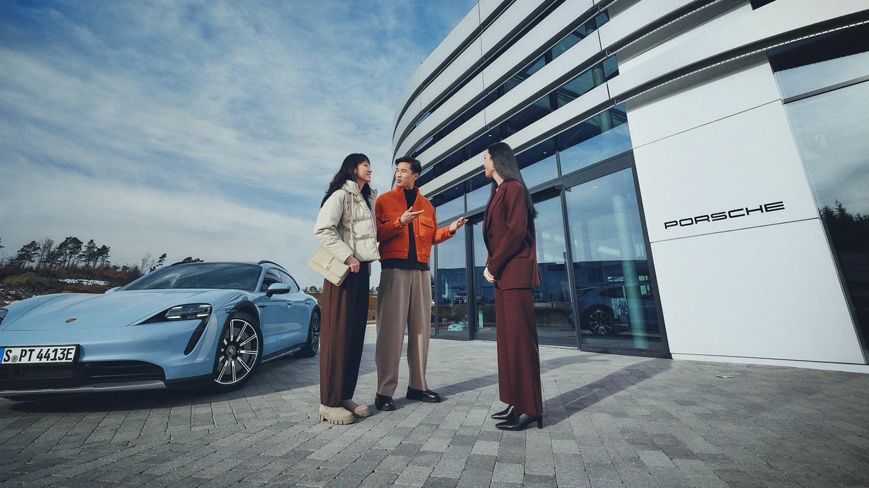 在保时捷中心的大楼前，一位女士正在接待一男一女两位顾客，他们的身后停着一辆蓝色的Taycan。