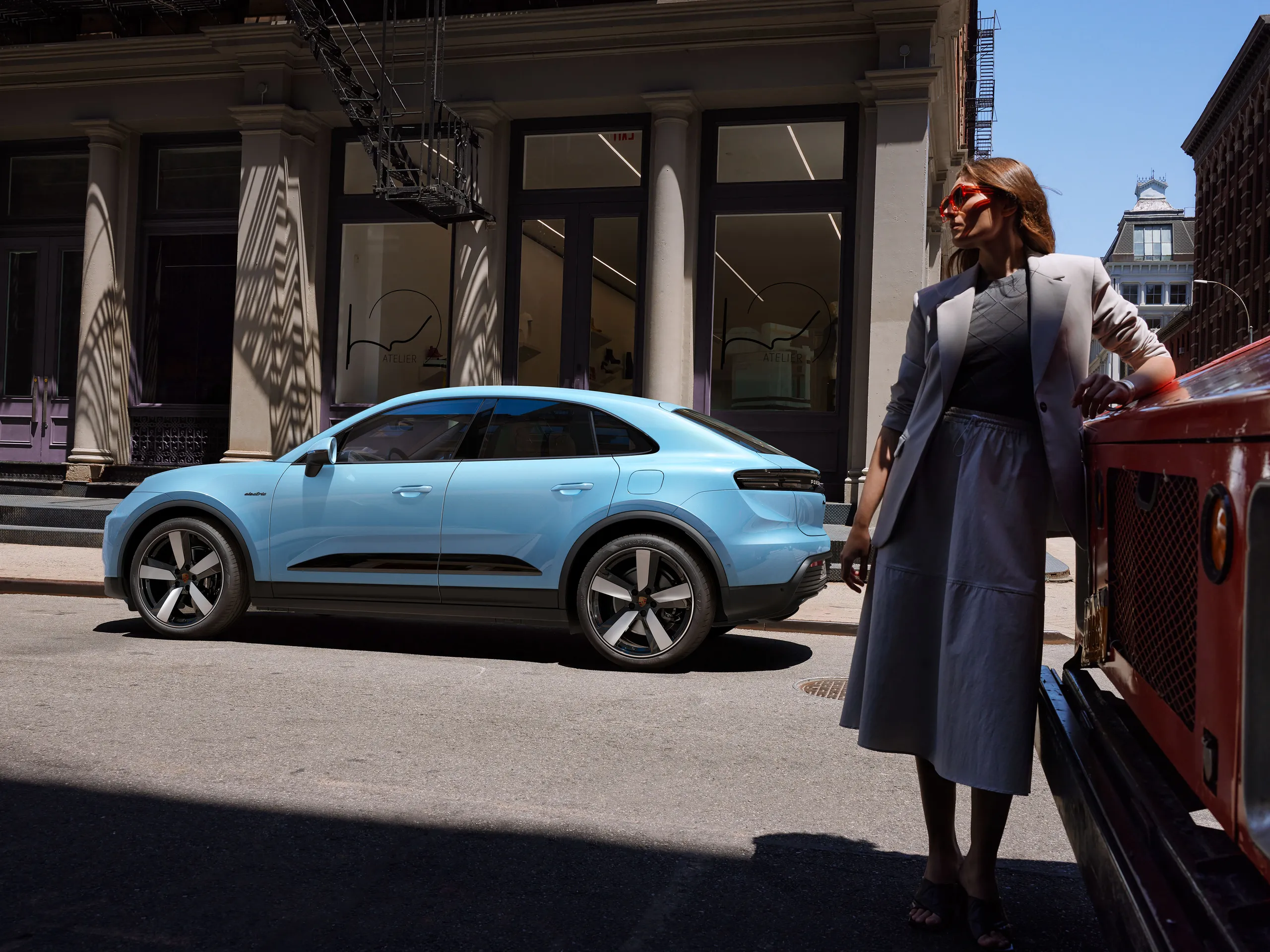 Side shot of a Frozen Blue Metallic Macan Electric parking in front of a building in the city with a woman standing next to it.