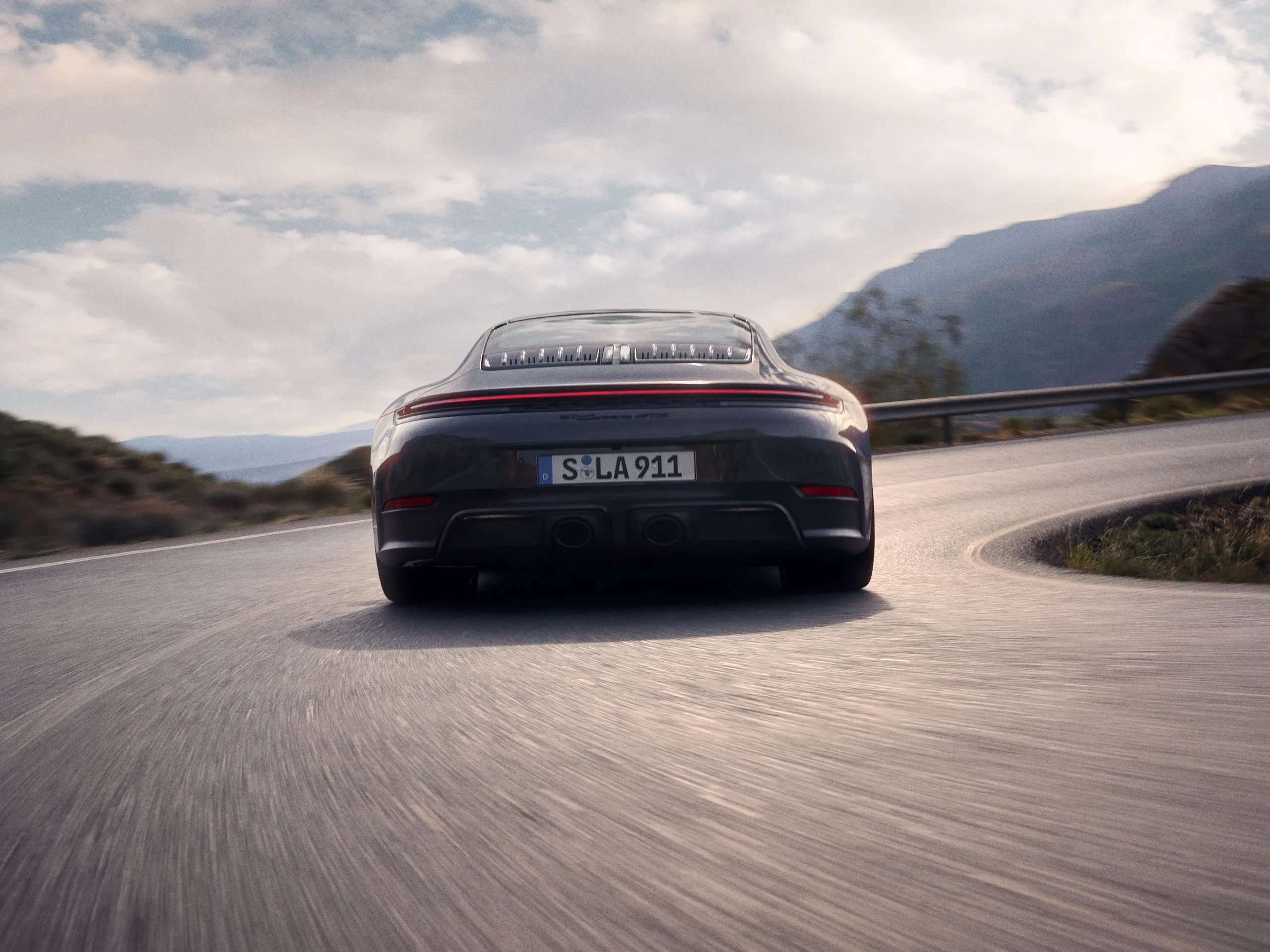 Rear shot of  a Slate Grey Neo colored 911 Carrera GTS driving on a street in the mountains.